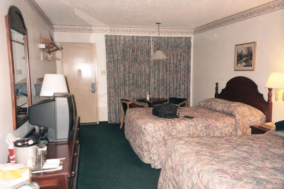 A dated motel room with made beds, a patterned bedspread, and heavy curtains drawn against a parking lot.